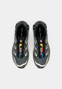 Fekete atlétikai cipő texturált felsőrésszel, fehér díszítésekkel, sárga, piros és kék csíkokkal, valamint fűzőkkel a biztos illeszkedésért.