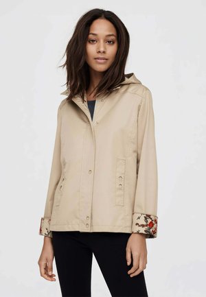 Donna che indossa una giacca beige con cappuccio e polsini stampati con motivi floreali, abbinata a pantaloni neri, in piedi davanti a uno sfondo neutro.