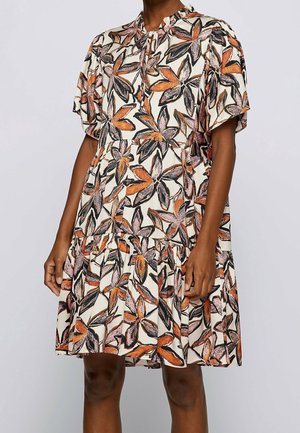 Femme portant une robe ample à manches courtes avec un col à volants et un motif floral orange, noir et rose sur fond crème.