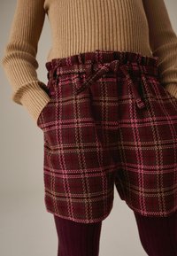 Pantaloni corti a quadri in bordeaux con righe rosa e beige. Presentano una vita arricciata con un laccio e due tasche frontali. Tessuto testurizzato.