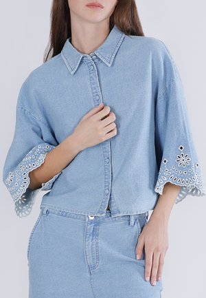 Vrouw draagt een lichtblauwe denim outfit met een overhemd met knopen en brede mouwen, afgewerkt met witte geborduurde kanten rand, en een bijpassende rok.