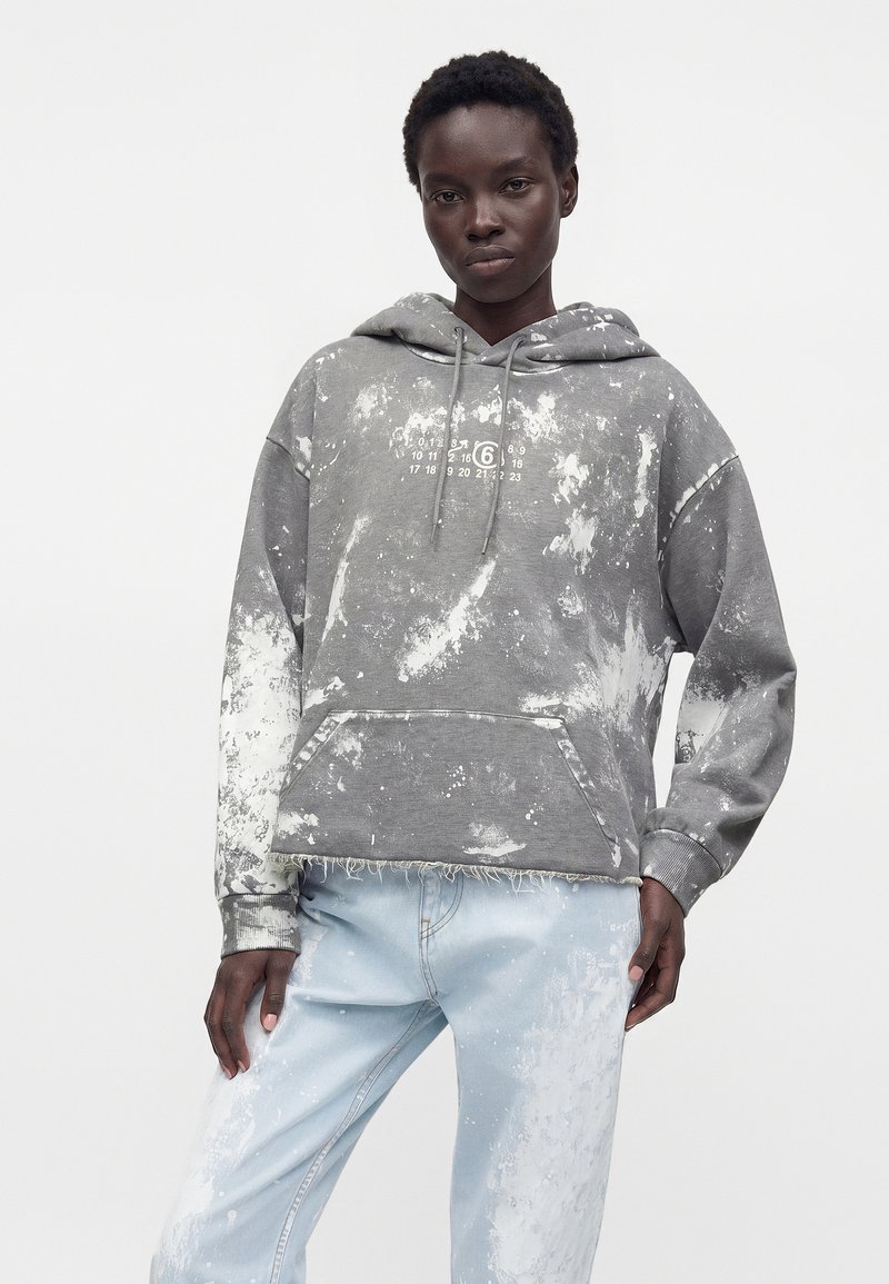 Person wearing a gray tie-dye hoodie with a calendar print and light blue jeans with white paint splatters, standing against a plain background.