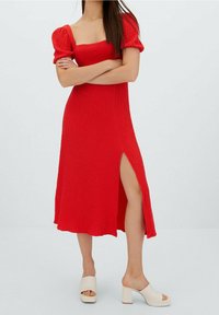 Czerwona sukienka do kolan z kwadratowym dekoltem i bufkami. Teksturowany materiał z bocznym rozcięciem. Noszona z białymi sandałami na słupku.
