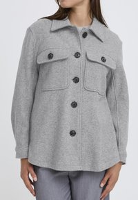 Šedý vlnený kabát so štyrmi vreckami, čiernymi gombíkmi a špicatým golierom. Tkanina má jemný, textúrovaný povrch. Skombinované so šedými nohavicami.