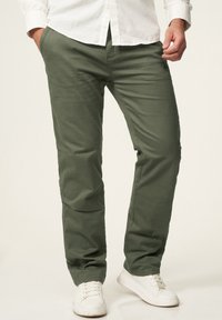 Green cotton pants with a straight leg, front pockets, and slight sheen. Paired with white sneakers for a casual look.