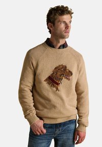 Beezikas spordisärk, millel on kudumise tekstuur, rohelise koera pea embleem pruunis, punase kaelarihmaga detailiga ja soonitud varrukate ning äärega.