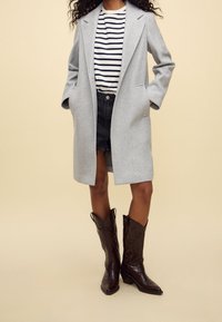 Gray wool blend coat with wide lapels, paired with a striped shirt and frayed black denim shorts, complemented by brown leather cowboy boots.