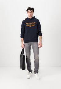 Marineblauwe hoodie met oranje tekst, grijze jeans, witte sneakers en een zwarte totebag. De nadruk ligt op eenvoudig ontwerp en functionele kleding.