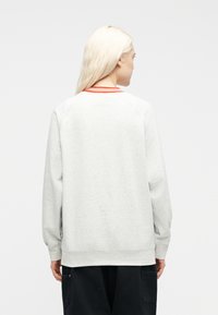 Άτομο με μακριά ξα νθά μαλλιά που φοράει ανοιχτό γκρι φούτερ με πορτοκαλί ριγέ γιακά, στέκεται με την πλάτη προς το φακό μπροστά σε λευκό απλό φόντο.