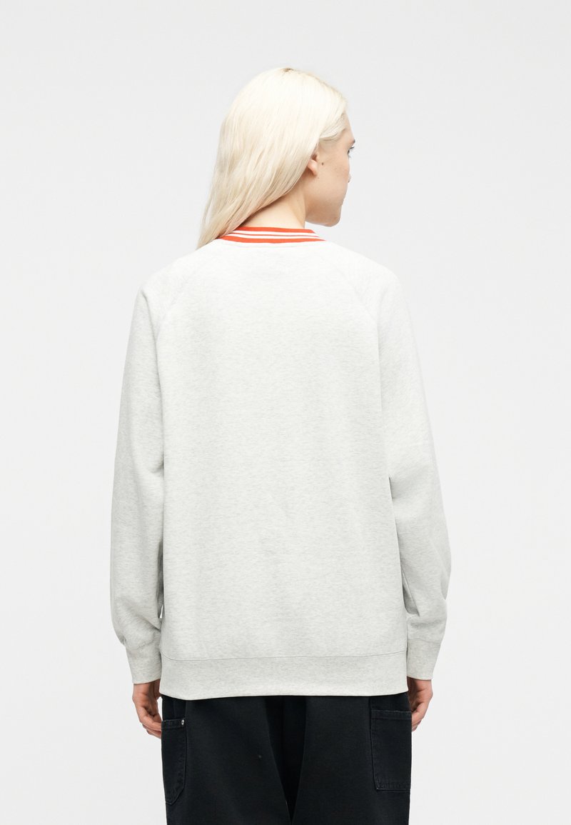 Person mit langen blonden Haaren, der einen hellgrauen Sweatshirt mit orange gestreiftem Kragen trägt und mit dem Rücken zu einer schlichten weißen Wand steht.