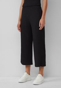 Schwarze, geschnittene weite Hosen aus strukturiertem Stoff mit elastischem Bund, kombiniert mit weißen Sneakers vor einem neutralen Hintergrund.
