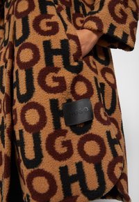 Brauner Fleece-Mantel mit großen schwarzen und burgunderroten Buchstaben, die "HUGO" schreiben. Mit einem Logopatch, abgerundetem Saum und Seitentaschen. Weicher Stoff.
