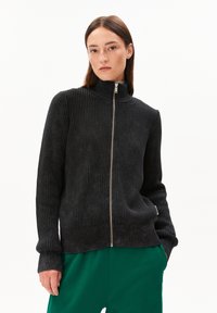 Schwarzer, gerippter Zip-Up-Pullover mit hohem Kragen, strukturiertem Stoff, taillierter Silhouette und kontrastierenden grünen Hosen. Einfaches Design, keine Muster.
