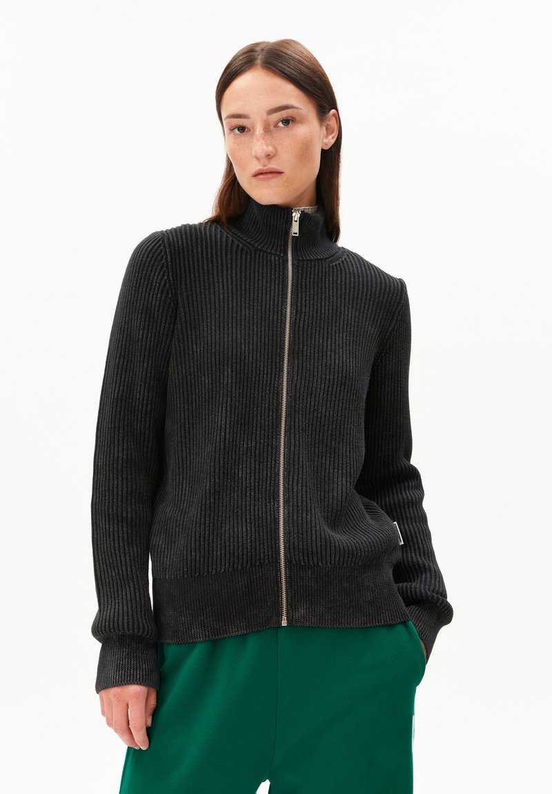 Schwarzer, gerippter Zip-Up-Pullover mit hohem Kragen, strukturiertem Stoff, taillierter Silhouette und kontrastierenden grünen Hosen. Einfaches Design, keine Muster.