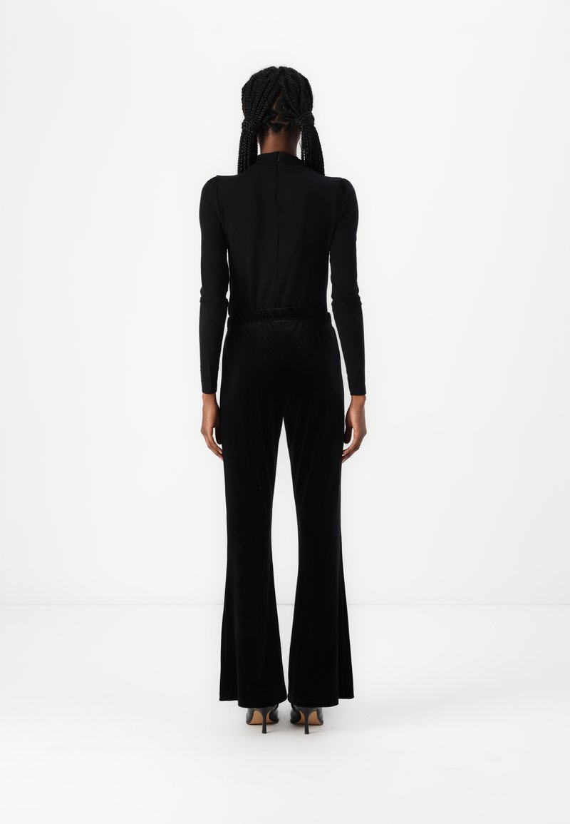 Top nero aderente con maniche lunghe e collo alto, abbinato a pantaloni svasati in velluto nero e scarpe con tacco alto. Sfondo semplice.