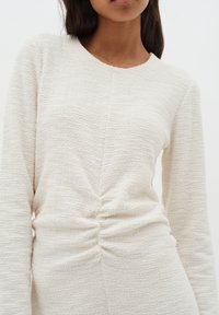 Vit texturerad långärmad topp med rund halsringning och samlad detalj vid midjan. Lätt, mjukt tyg med en subtil, ojämn yta.