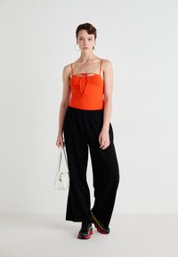 Orange fitted tank top with thin straps, black wide-leg pants, holding a white handbag, and wearing black and red sneakers.