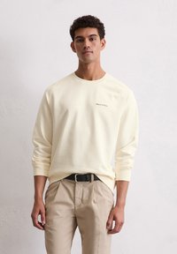Hanorac galben deschis cu guler din tricot și mâneci lungi, având un logo mic pe pieptul stâng. Asortat cu pantaloni bej.