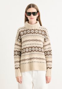 Pull en tricot beige avec un col montant, des bords côtelés et des motifs géométriques marron distincts sur un fond crème.