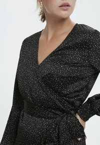 Vestido negro envolvente de tejido suave con lunares blancos, que presenta mangas largas y un detalle de lazo fruncido en la cintura.