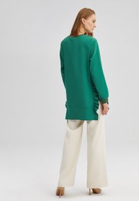 Top verde a maniche lunghe con arricciatura laterale, abbinato a pantaloni bianchi a gamba larga. Il top presenta una vestibilità comoda e un orlo arrotondato.
