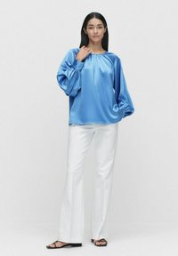 Satinblå blus med puffärmar och rynkad halsringning. Matchas med vita utsvängda byxor och svarta platta sandaler. Modellen står mot en enfärgad bakgrund.