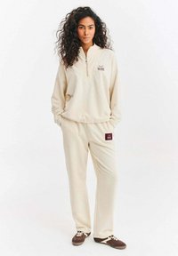 Béžový fleecový pulovr s límcem na zip, doplněný odpovídajícími tepláky s logovou nášivkou. Texturovaný, volný střih.