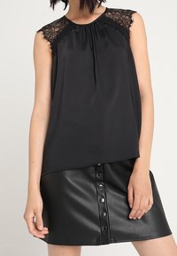 Svart ärmlös topp med spetsdetaljer vid axlarna, plisserad front, parat med en svart fusk läderkjol med knappstängning.
