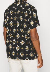 Mężczyzna ubrany w czarną koszulę z krótkim rękawem w żółte i białe geometrczne wzory, w połączeniu z białymi spodniami, pokazany od tyłu.