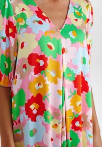 Flerfärgad blus med blommig design i lätt tyg. Har en V-ringning och korta puffärmar. Dominerande rosa med röda, gröna, gula och blå blommor.