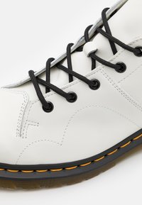 Zapato de cuero blanco con cordones negros, suela de goma, costuras amarillas y punta redondeada. Presenta una textura suave y un sutil detalle en la curvatura.