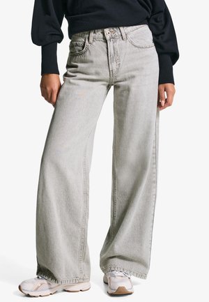 Osoba oblečená v svetlosivých širokých džínsoch, bielych teniskách a čiernom tričku s dlhým rukávom a rebrovanými manžetami, stojaca na bielom pozadí.