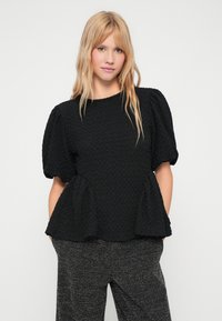 Top preto texturizado com mangas curtas bufantes, decote redondo e bainha peplum. Usado com calças escuras estampadas.
