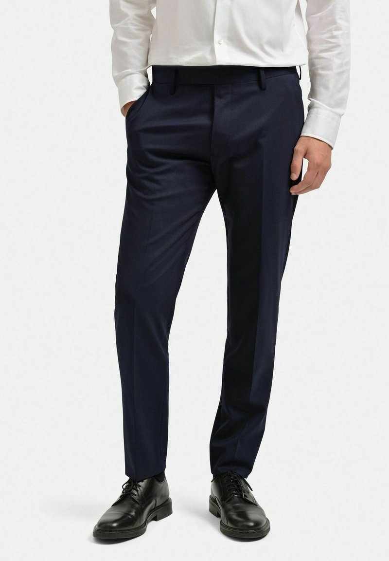 Marinblå byxor i skräddarsydd passform, tillverkade av mjukt tyg, med slät front, sidofickor och bälteshällor. Bärs med svarta skor.