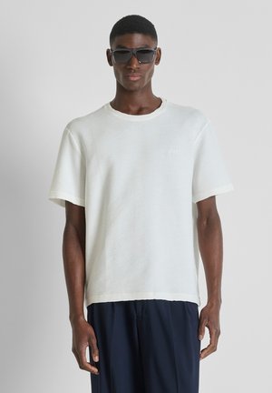 Man met zwarte zonnebril, wit textuuroverhemd met korte mouwen en donkere broek, staand tegen een effen grijze achtergrond.