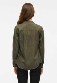Camicia verde oliva a maniche lunghe realizzata in tessuto morbido, con orlo curvo e polsini con bottoni, mostrata di spalle con pantaloni neri.
