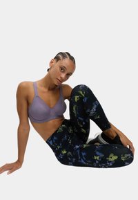 Paarse sportbeha met gaasdetails en zwarte legging met een kleurrijk bloemenpatroon. Model poseert zittend met zwarte sportschoenen.