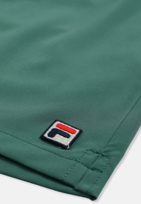 Tissu vert avec un patch logo Fila cousu en bleu marine, rouge et blanc, près d'un ourlet et d'une couture.