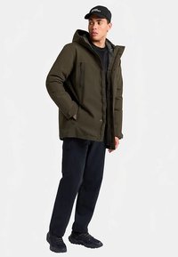 Olivgrüne, wattierte Jacke mit Reißverschluss und Druckknopfverschluss, großer Kapuze, zwei Reißverschlusstaschen, getragen über einem schwarzen Hemd und dunkler Hose.