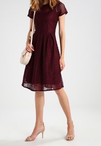 Burgunderfarbenes Spitzenkleid mit kurzen Ärmeln, tailliertem Oberteil und ausgestellt Rock; kombiniert mit einer hellrosa Umhängetasche und nudefarbenen High Heels.
