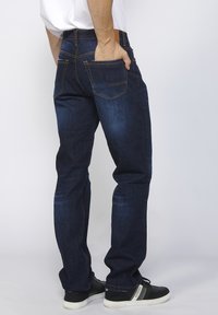 Donkerblauwe denim jeans met een rechte pasvorm, voorzien van achterzakken en contrasterende gele stiksels. G gedragen met zwarte sneakers.