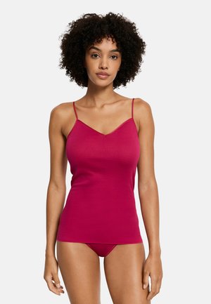 Femme aux cheveux bouclés portant un caraco rose vif ajusté et des sous-vêtements assortis, debout devant un fond blanc uni.