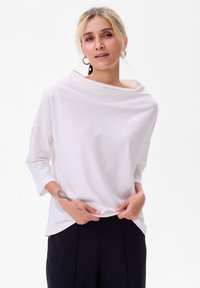 Witte lange mouwen top met een bootnek, gemaakt van zachte stof, met een ontspannen pasvorm en een licht gedrapeerd ontwerp. Gecombineerd met donkere broek.