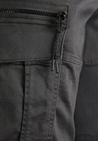 Černá látka s hladkým povrchem, obsahující viditelný zip a dvojité táhla, prošívané detaily a strukturovanou kapsu.