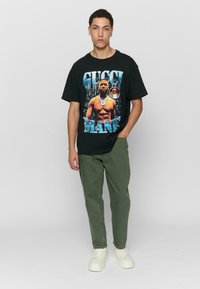 Black graphic t-shirt featuring a large print of a muscular figure and text, paired with olive green pants and white sneakers. Plain background.