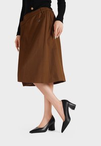Tacchi a punta in pelle nera con tacco largo, abbinati a una gonna plissettata marrone con tasca laterale e dettaglio dei bottoni.