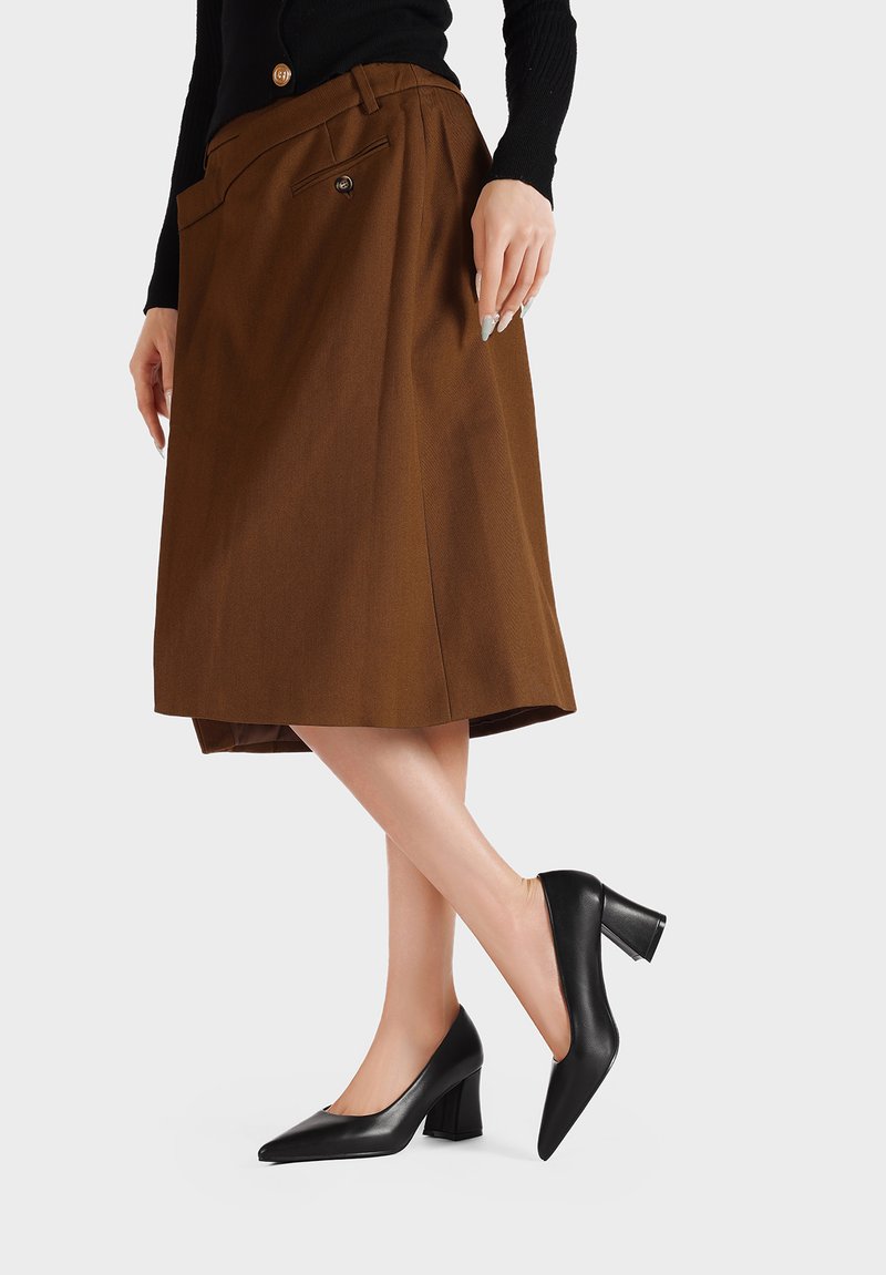 Tacchi a punta in pelle nera con tacco largo, abbinati a una gonna plissettata marrone con tasca laterale e dettaglio dei bottoni.