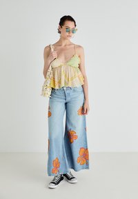 Lys grønn og gul mønstrete peplum-top med volangkant, sammen med vide lys blå jeans med oransje blomstertrykk. Svarte joggesko.