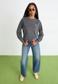 Woman with long braids wearing gray long-sleeve shirt, blue jeans, beige sneakers, and sunglasses standing on green and white floor.