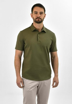 Man staat voor een effen achtergrond, draagt een olijfgroen poloshirt met korte mouwen en lichtbeige broek, kijkt neutraal vooruit.